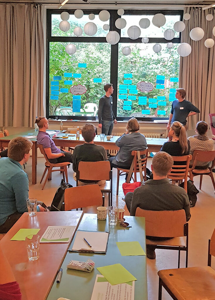 Mehrere Teilnehmer:innen an Tischen sitzend, zwei Vortragende stehend vor einem Fenster mit bunten Zetteln, Eindrücke vom Networking-Camp Science 1.0 des HCCI, Institut für Supervision in Berlin