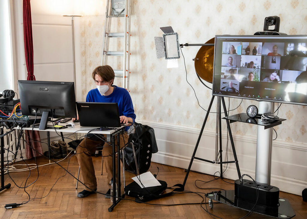 Mann mit Mundschutz an einem Tisch mit Computer- und Aufzeichnungstechnik sitzend, links davon ein Monitor mit Online-Meeting; Eindrücke vom Networking-Camp Science 2.0 des HCCI, Institut für Supervision in Berlin